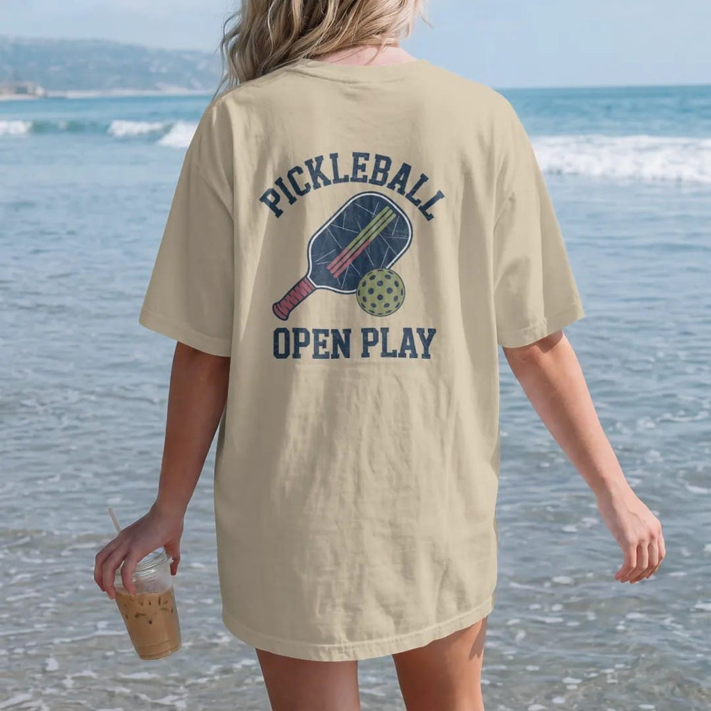 Woman on the beach in a Sandstone tee, back view: a paddle and a holed plastic ball with speed lines, with "PICKLEBALL" and "OPEN PLAY" in chunky faded collegiate block letters, printed in faded varsity navy, washed red, mustard gold and pine green…
