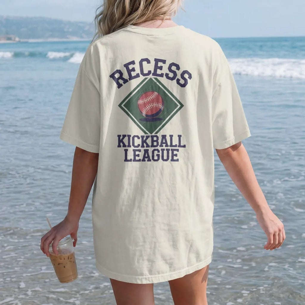 Woman on the beach in a Ivory tee, back view: a bouncing red kickball over a chalk-drawn diamond, with "RECESS" and "KICKBALL LEAGUE" in chunky rounded 70s block letters, printed in faded varsity navy, washed red, mustard gold and pine green with a…