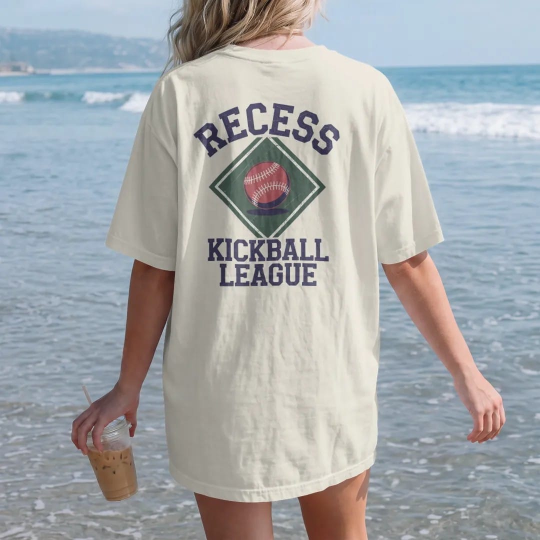 Woman on the beach in a Ivory tee, back view: a bouncing red kickball over a chalk-drawn diamond, with "RECESS" and "KICKBALL LEAGUE" in chunky rounded 70s block letters, printed in faded varsity navy, washed red, mustard gold and pine green with a…