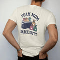 Man wearing a Ivory tee, seen from the back: a cooler of orange slices with juice boxes and a stack of snack bags with stars, with "TEAM MOM" and "SNACK DUTY" in chunky 1970s swash script, printed in faded varsity navy, washed red, mustard gold and…