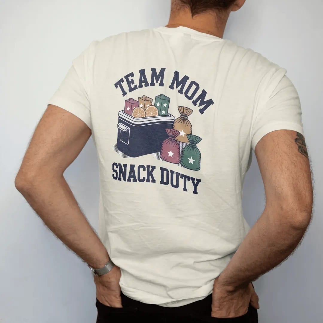 Man wearing a Ivory tee, seen from the back: a cooler of orange slices with juice boxes and a stack of snack bags with stars, with "TEAM MOM" and "SNACK DUTY" in chunky 1970s swash script, printed in faded varsity navy, washed red, mustard gold and…