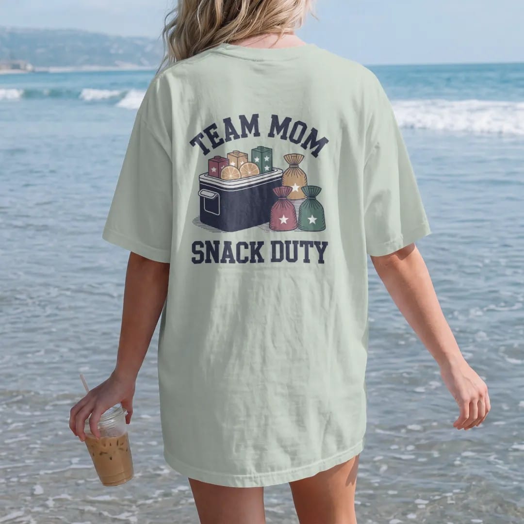 Woman on the beach in a Bay tee, back view: a cooler of orange slices with juice boxes and a stack of snack bags with stars, with "TEAM MOM" and "SNACK DUTY" in chunky 1970s swash script, printed in faded varsity navy, washed red, mustard gold and…