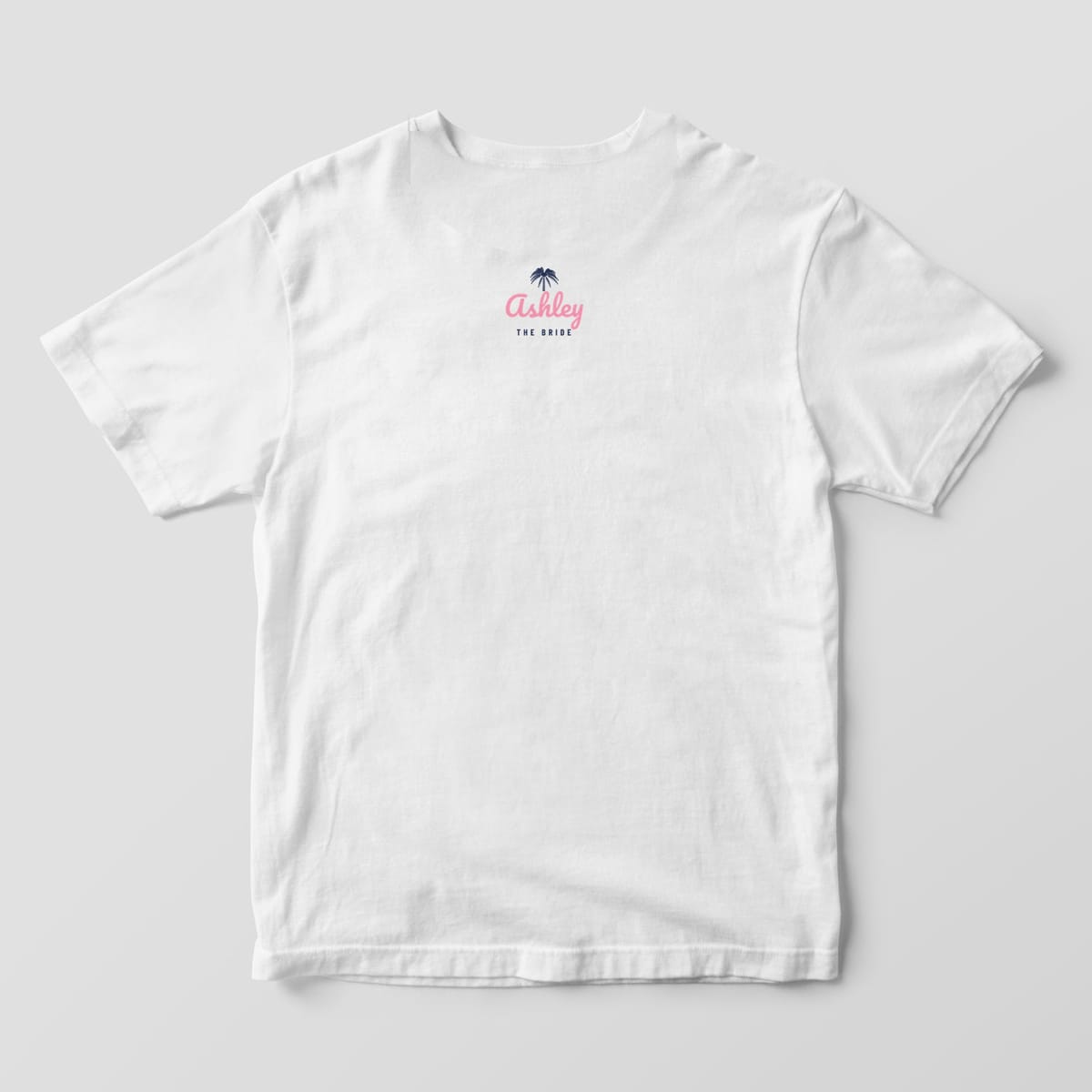 Back of a white Comfort Colors 1717 tee with a small navy palmetto, Ashley in pink script and THE BRIDE below the collar. A BRIDE'S CREW version is offered.