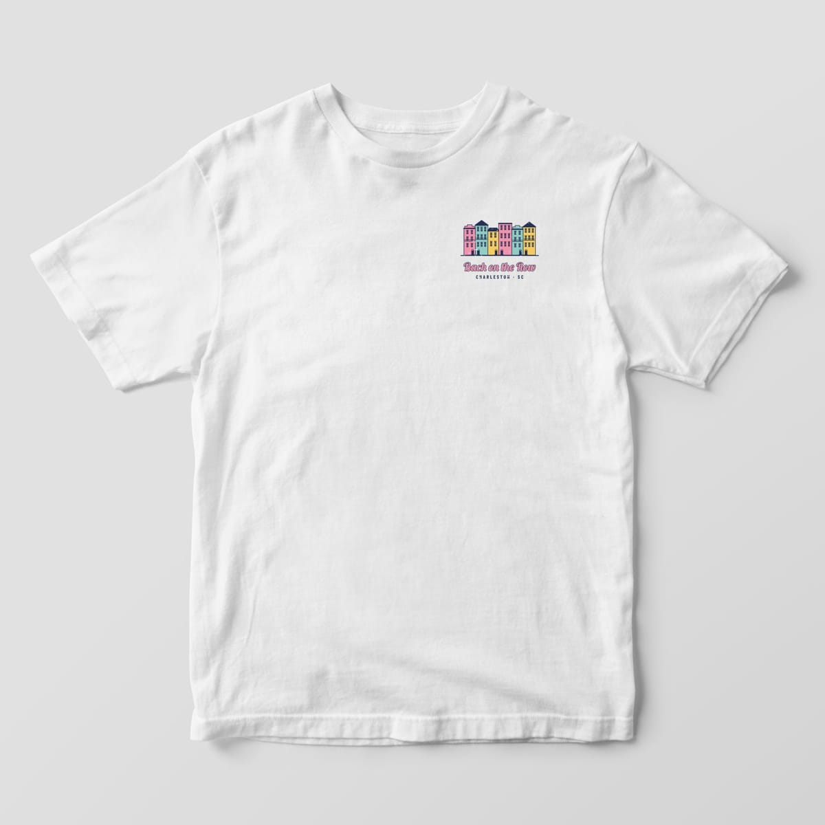 Front of a white Comfort Colors 1717 tee with a small pastel row house chest print and Bach on the Row script.