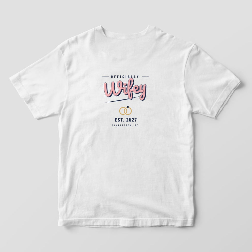 Back of a White Comfort Colors tee: OFFICIALLY above pink retro Wifey script with navy shadow, gold rings, EST. 2027 and CHARLESTON, SC