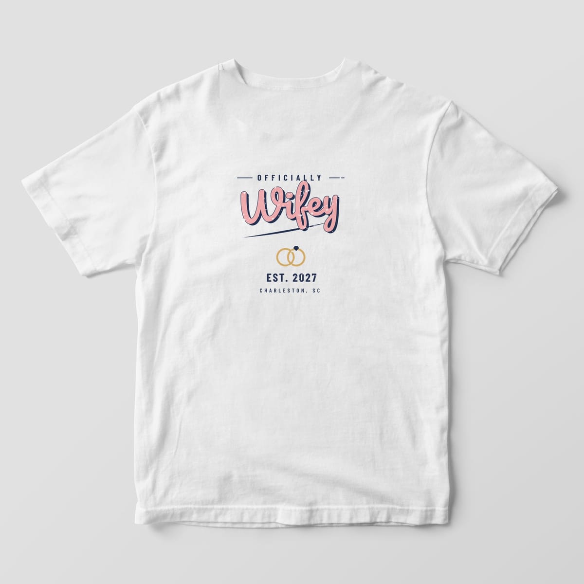 Back of a White Comfort Colors tee: OFFICIALLY above pink retro Wifey script with navy shadow, gold rings, EST. 2027 and CHARLESTON, SC